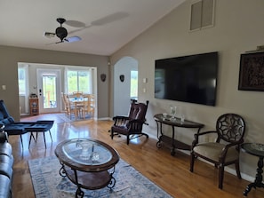 A spacious living room with a ceiling fan, a large flat-screen TV, a leather couch, a glass coffee table, and a dining area with chairs and a table.