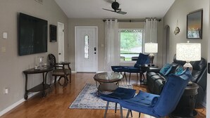 Ein geräumiges Wohnzimmer mit Holzdielen, einem Deckenventilator, einem großen Flachbildfernseher, einem Couchtisch und einer Sitzecke mit blau gepolsterten Sesseln und einem Sofa.