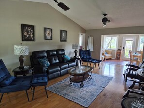 A living room with a black leather sofa, blue armchairs, a coffee table, and a rug. There are framed pictures on the wall, a lamp, and a ceiling fan.