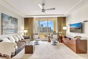 Una habitación de hotel con balcón, un sofá, sillones, un televisor y un cuadro.