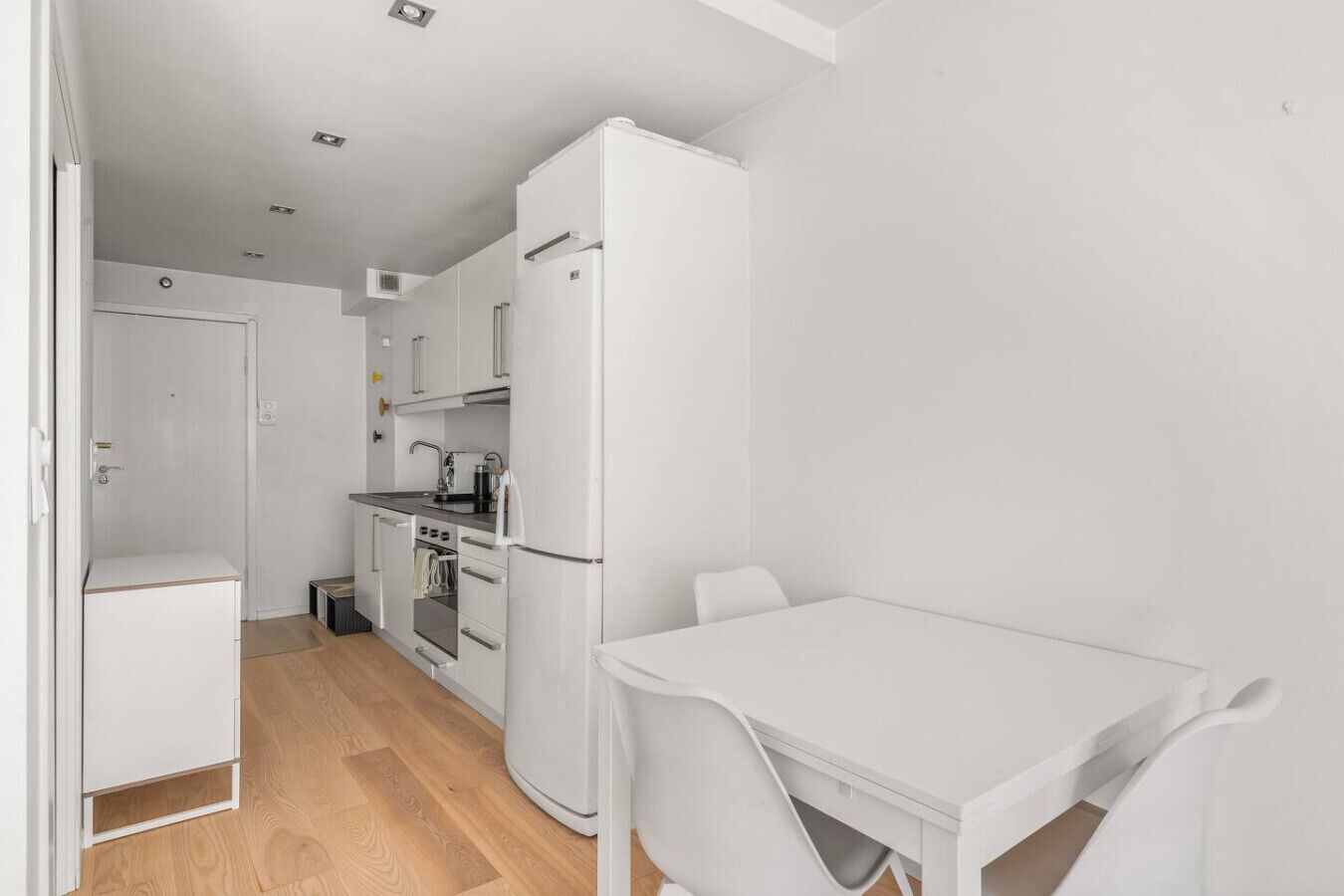 A compact kitchen with white appliances, a small oven, and a sink. There is a white table and chair set in the dining area.