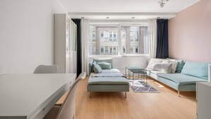 A modern, well-lit room with a bed, a sofa, a dining table, and a view of buildings through large windows.