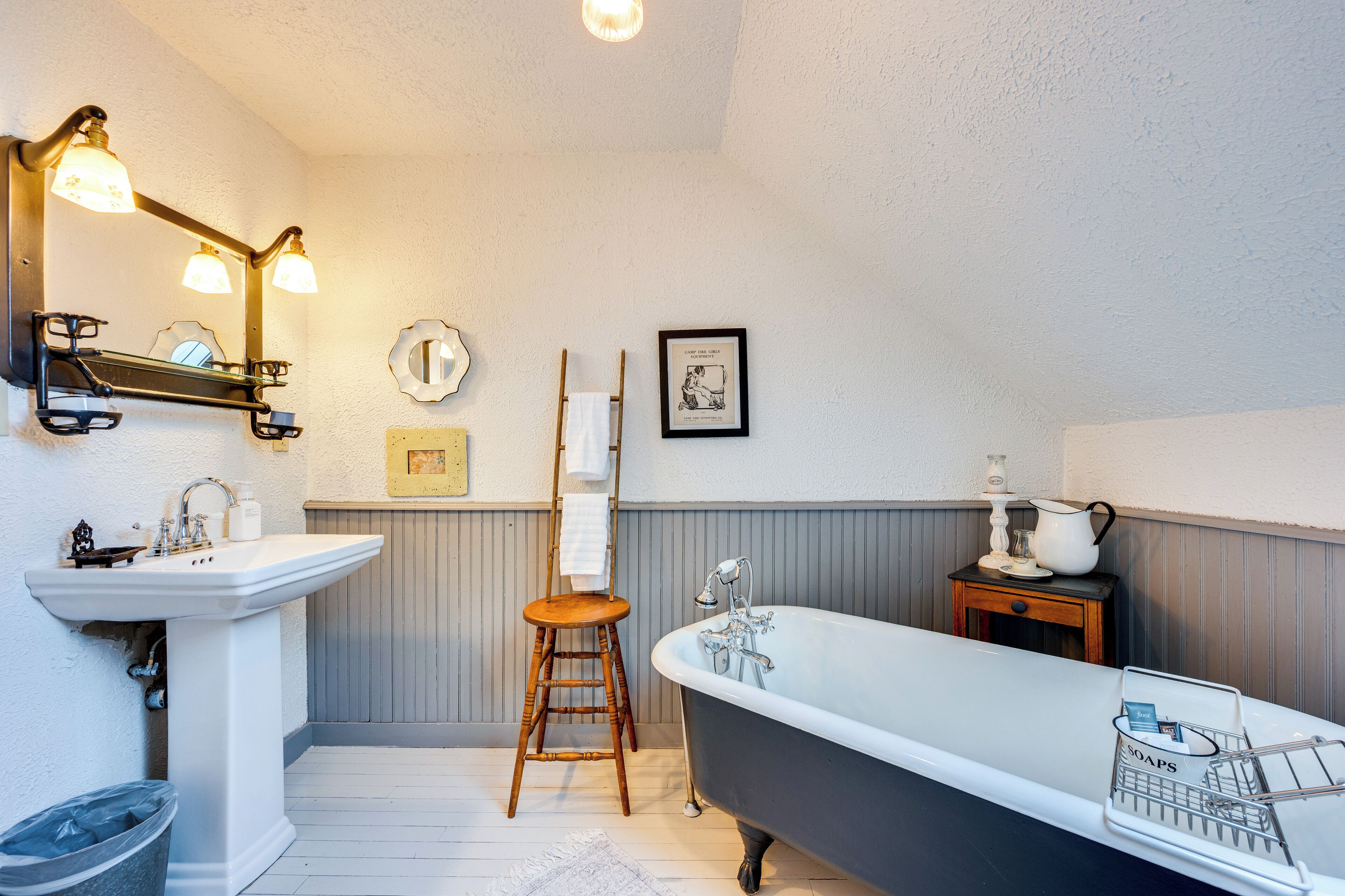 Ein Badezimmer mit einem Standwaschbecken, einer freistehenden Badewanne, einem hölzernen Hocker, einem Handtuchhalter und einem gerahmten Bild an der Wand.