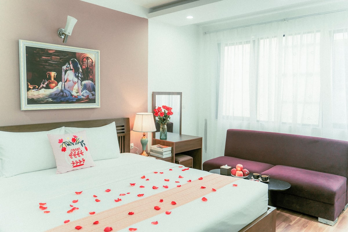 A hotel room with a bed, a sofa, a desk with a lamp, a vase with flowers, and a painting on the wall.