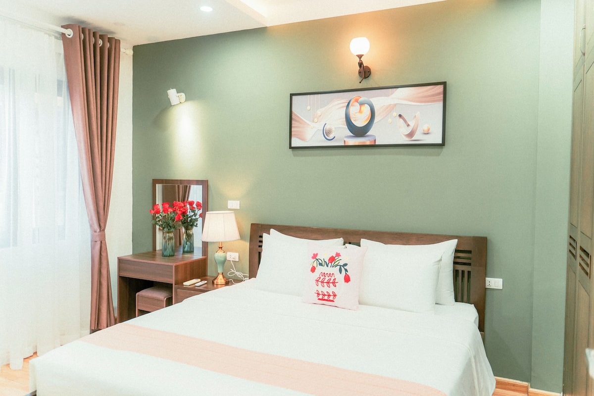 A hotel room with a bed, a nightstand, a lamp, a vase with flowers, and a painting on the wall.