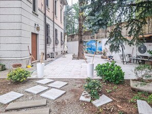 A courtyard with a tiled floor, potted plants, a tree, and a mural on the wall.