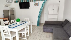 Un espacio habitable compacto con zona de comedor, un sofá gris, un refrigerador, un televisor montado en la pared y una escalera con detalles en color turquesa.