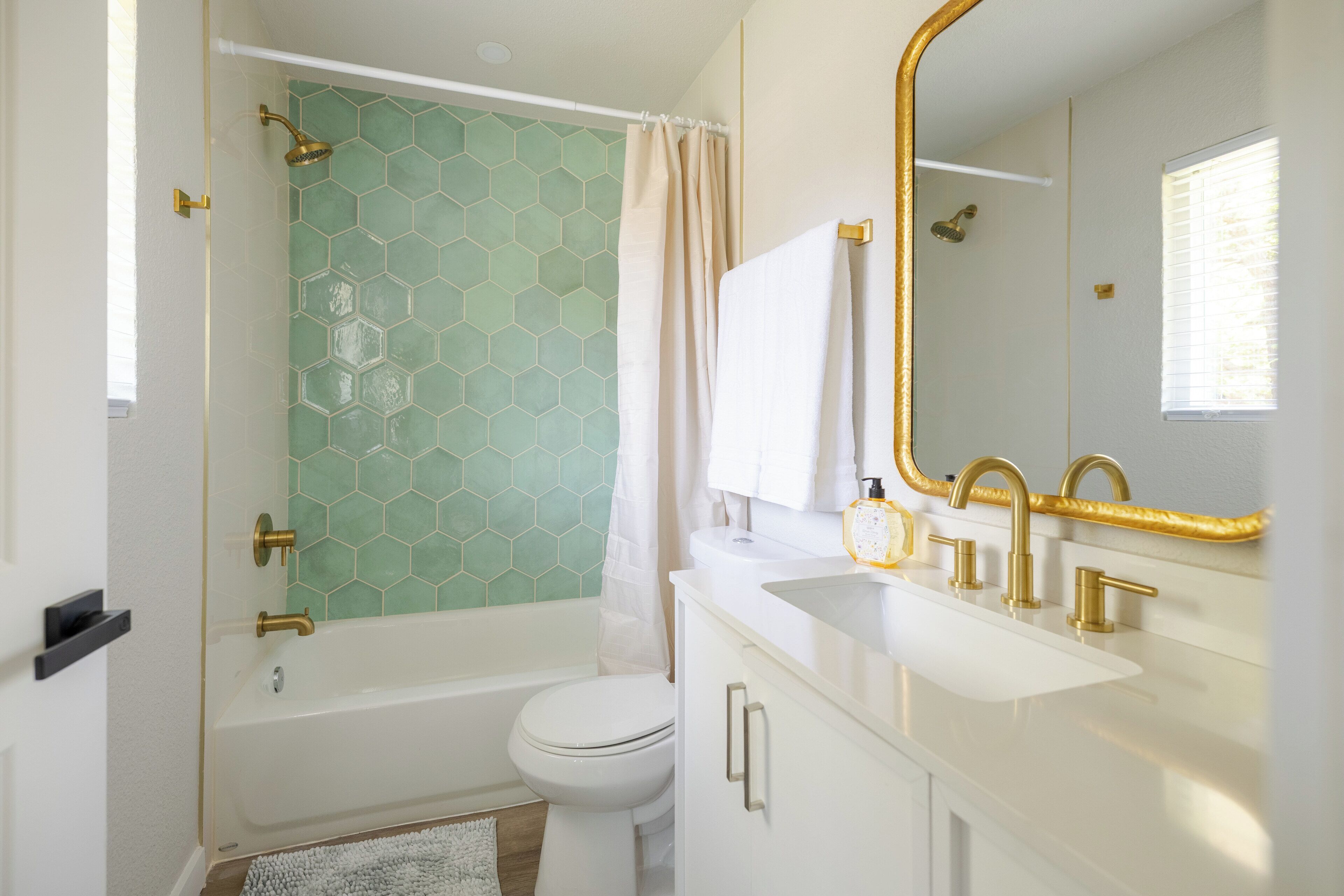 Een badkamer met een wand betegeld met groene hexagontegels, een witte badkuip, een wit toilet, een witte wastafel met gouden accenten, en een grote spiegel met gouden lijst.