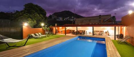Una piscina con cubierta de madera, césped y un edificio con tejado rojo.