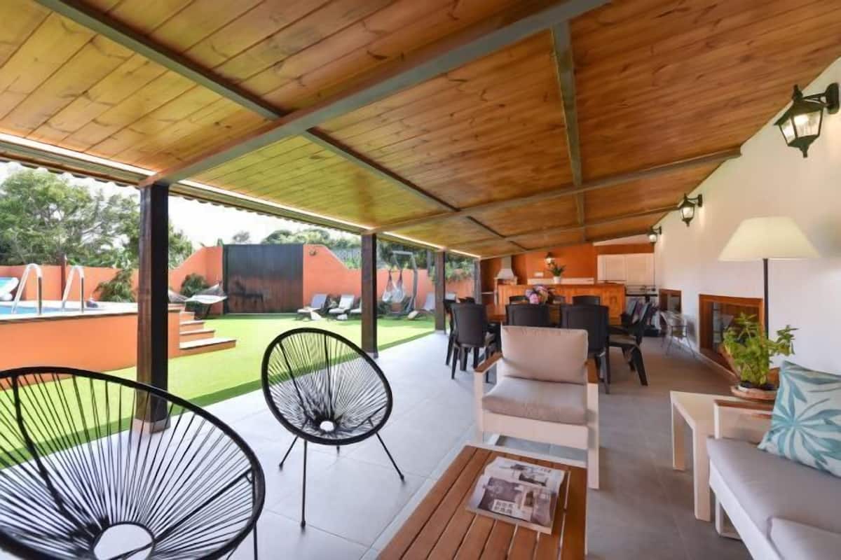 Un espacio al aire libre cubierto con techo de madera, zona de comedor y vistas a una piscina y jardín.