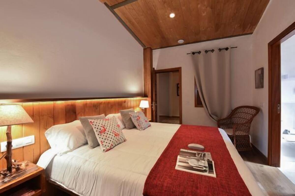 Quarto com teto de madeira, cama com roupa de cama branca e manta vermelha, mesa de cabeceira com abajur e cadeira de vime.