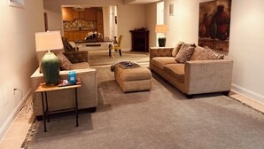 Ein Wohnzimmer mit einer Couch, Hocker und Couchtisch, ein Essbereich mit Tisch und Stühlen sowie eine Küche im Hintergrund.