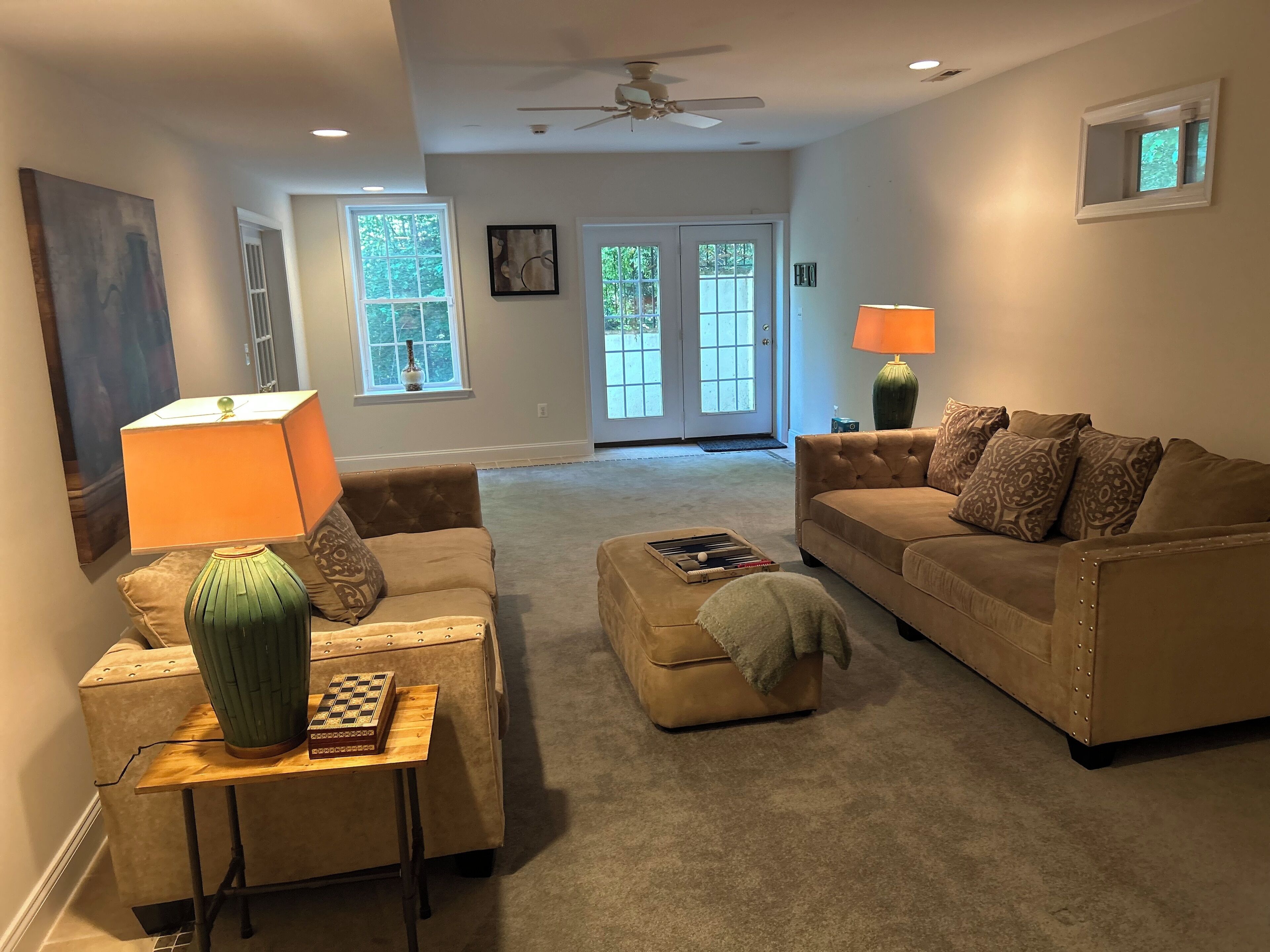 Sala de estar com dois sofás, uma mesa de centro, uma mesinha lateral com um tabuleiro de xadrez e um abajur. Há janelas com vista para árvores e uma porta que dá para o exterior.