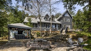 Une grande maison en bois avec une cheminée en pierre, des fauteuils Adirondack et une petite construction ornée d’enseignes.