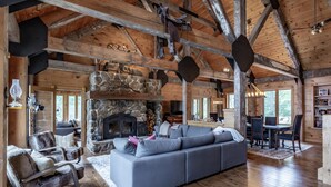 L’intérieur d’un chalet rustique, avec une cheminée en pierre, des poutres en bois, un canapé gris et un coin repas.