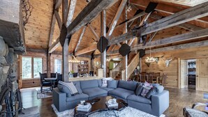 Un vaste salon doté d’un grand canapé d’angle gris, de poutres en bois au plafond, d’un coin repas avec des tables en bois et d’un escalier menant à un autre niveau.