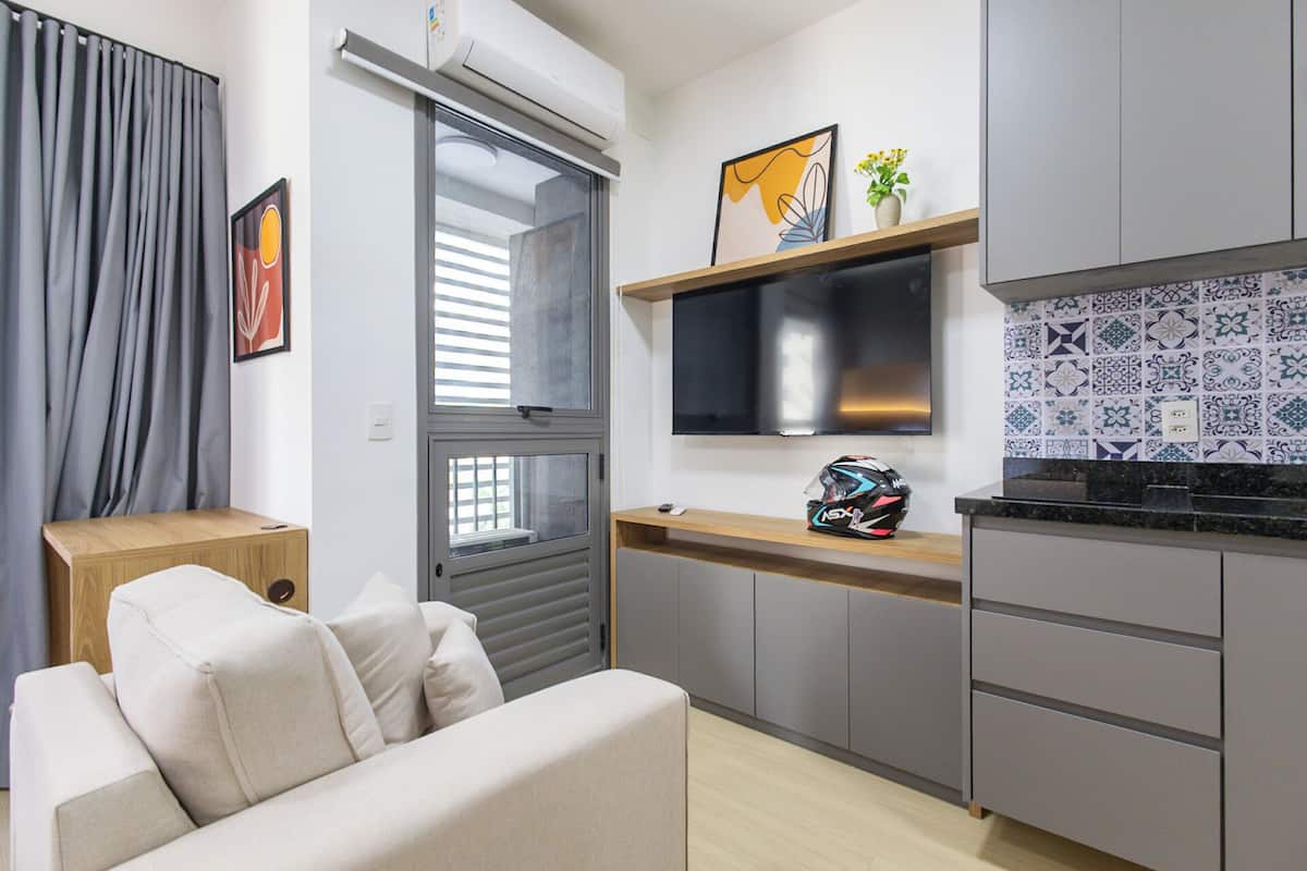 A modern kitchen with grey cabinets, a built-in oven, and a flat-screen TV. There is a sofa with cushions in the living area, a window with blinds, and a small plant on the counter.