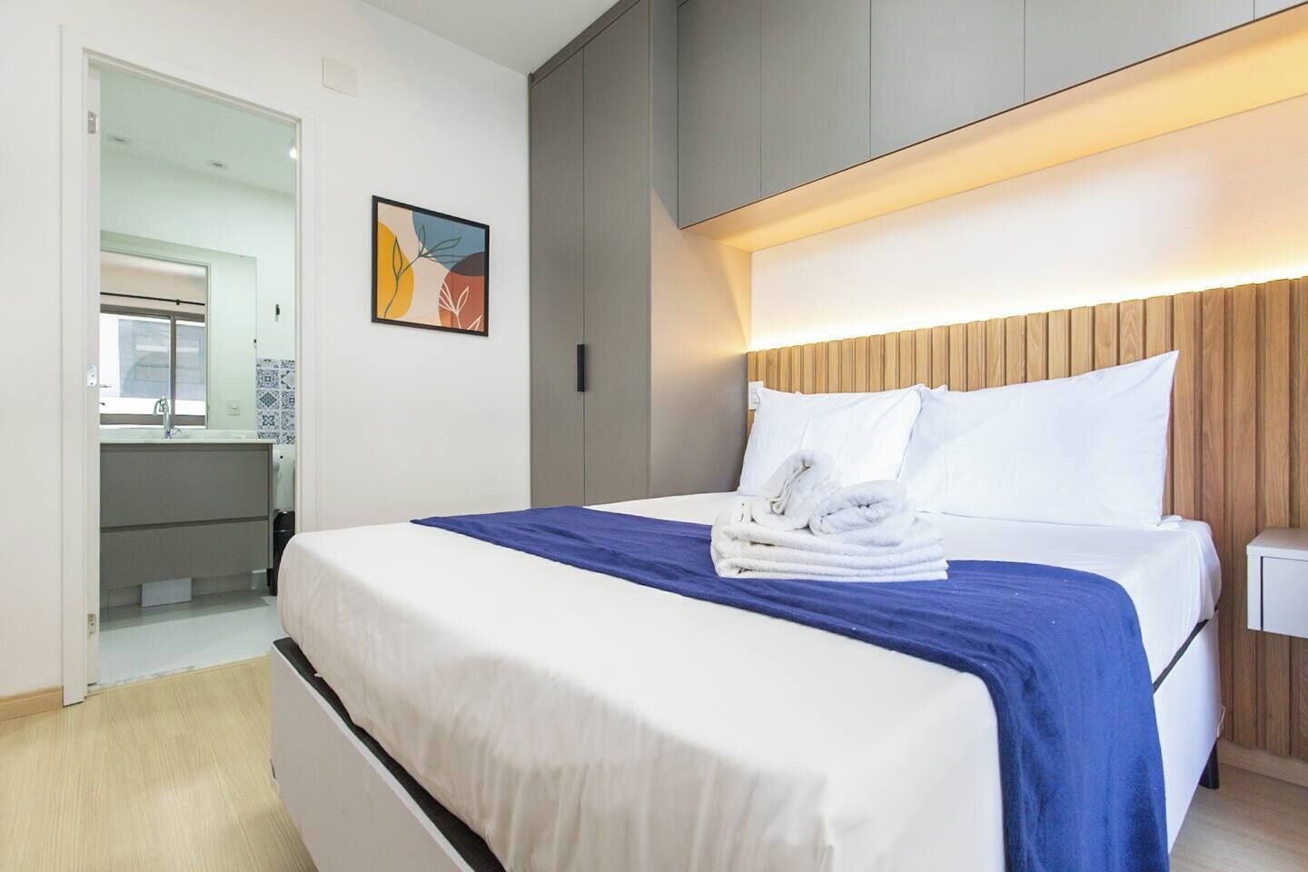 A modern hotel room with a large bed, a wooden headboard, a painting on the wall, and a bathroom visible through an open door.