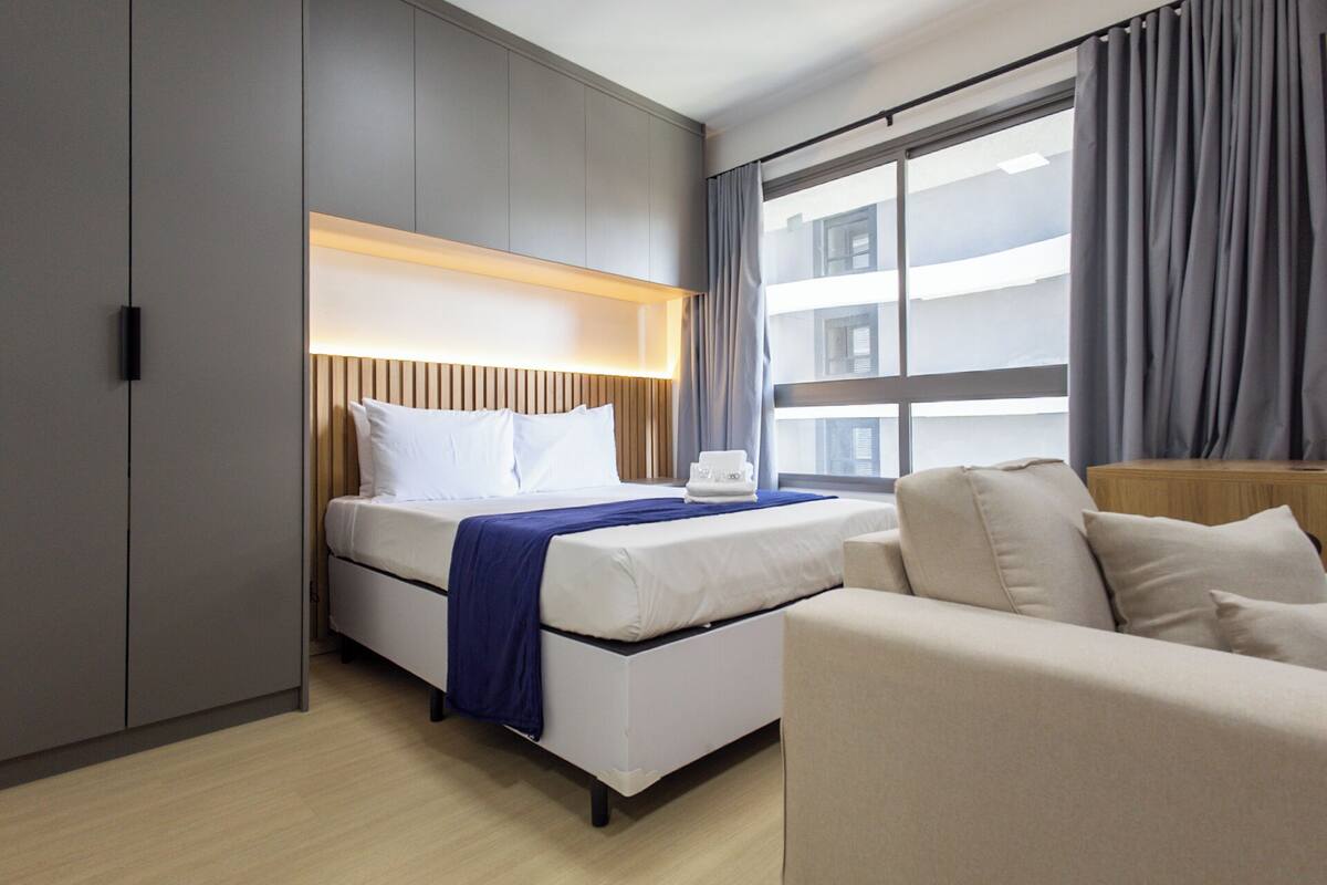 A hotel room with a bed, a sofa, a bedside table, a window with curtains, and a view of buildings outside.