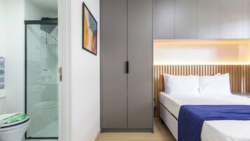 A hotel room with a shower, a bed with white bedding and a blue blanket, and a wooden headboard.