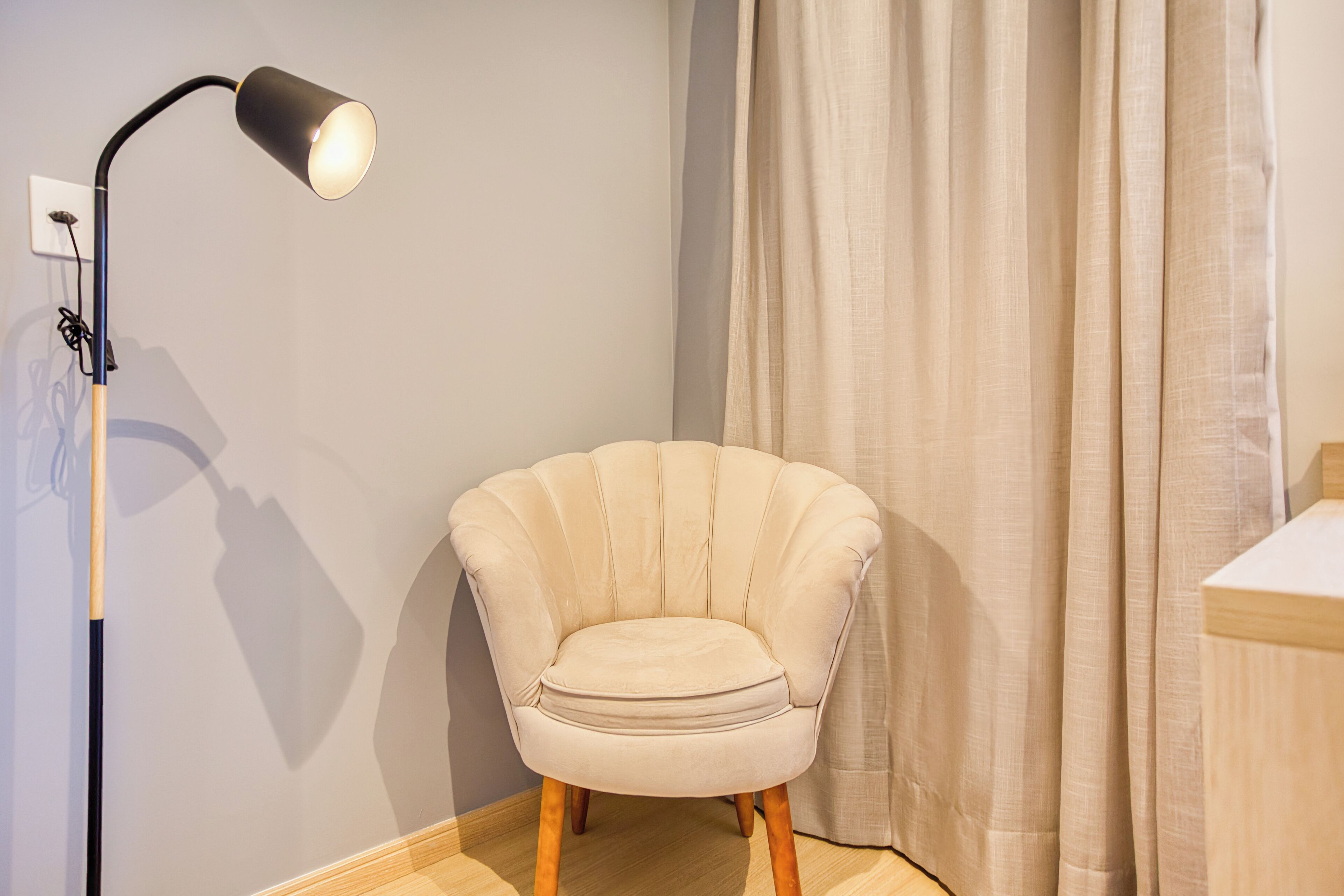 Une pièce avec un fauteuil beige, un lampadaire et un rideau.