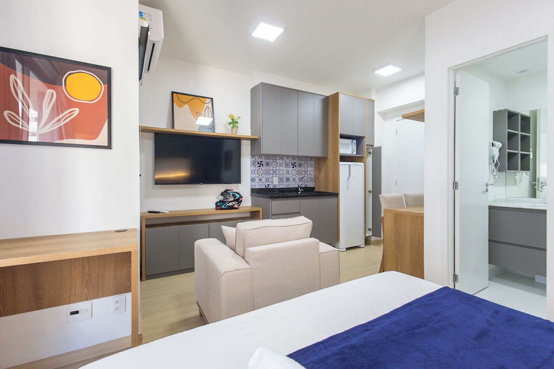 A compact hotel room with a bed, a desk with a chair, a kitchenette, and a TV.
