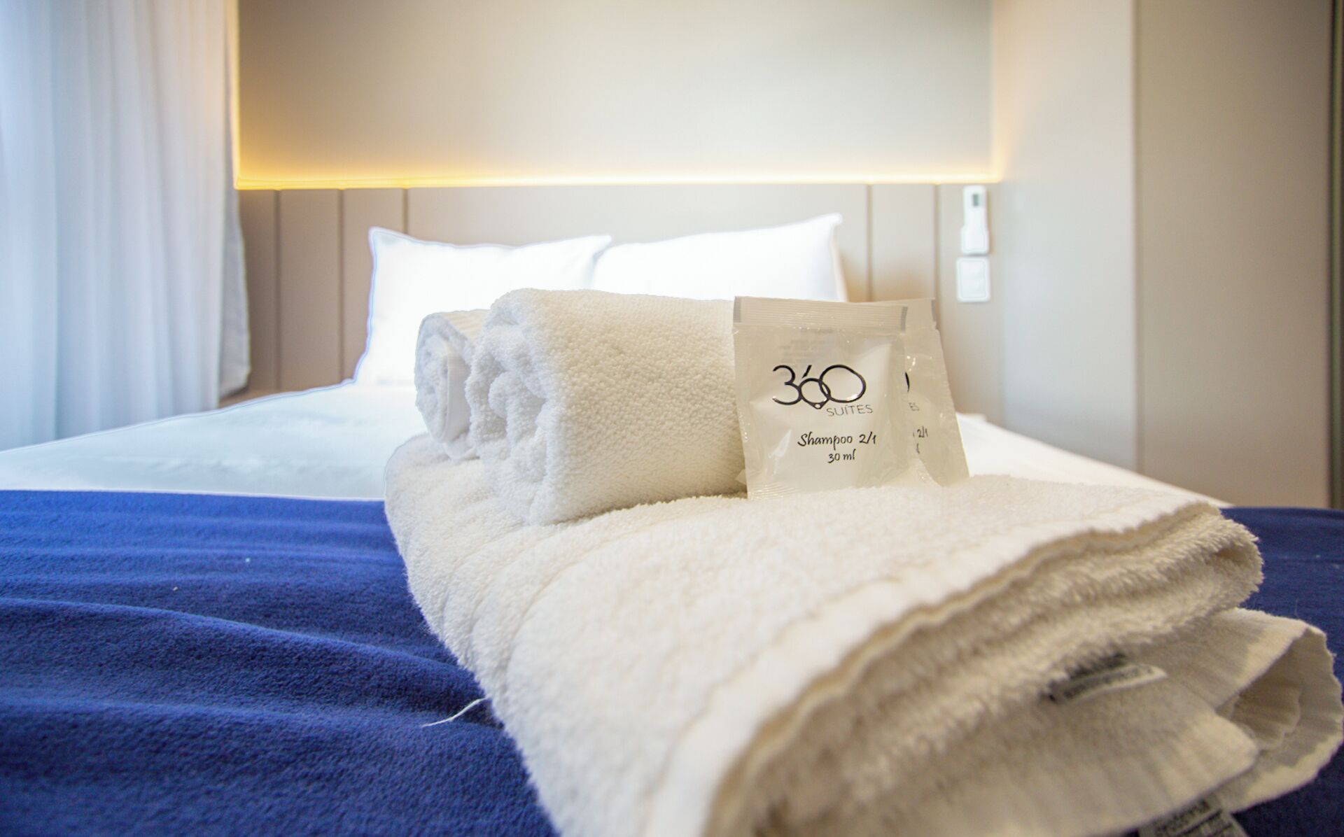 Cama bem arrumada com uma toalha branca dobrada e uma garrafa de shampoo.