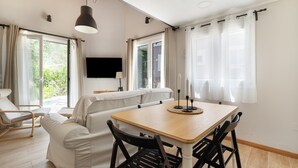 A modern living room with a wooden dining table, black chairs, a sofa, and a TV mounted on the wall.