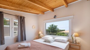 A bedroom with a large window, wooden ceiling beams, a bed with a pink blanket, two bedside lamps, and a view of greenery outside.
