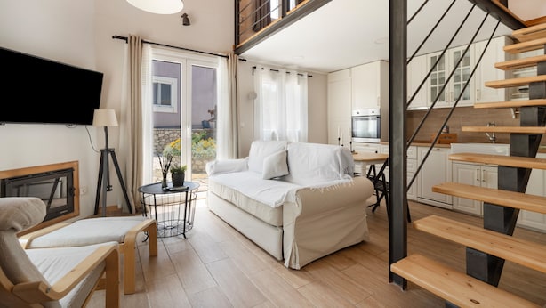 A modern living room with a sofa, a small table, a lamp, and a TV. There is a staircase leading to an upper level with a balcony.