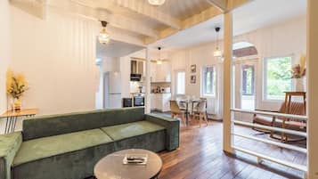 Een ruime woonkamer met een groene bank, een ronde salontafel en een eettafel met stoelen. Achterin is een keuken te zien, en de kamer heeft houten vloeren en grote ramen.