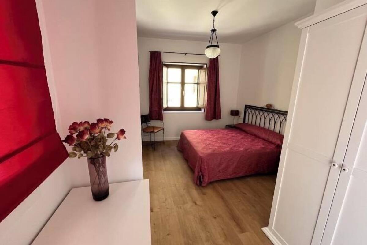A bedroom with a bed, a desk, a chair, a window with curtains, and a vase with flowers.