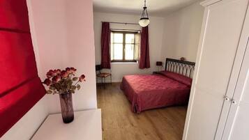 Un dormitorio con cama, escritorio, silla, ventana con cortinas y un florero con flores.