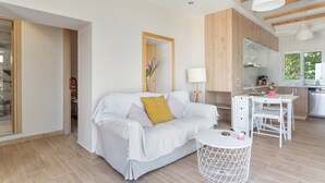 Un salon moderne avec un canapé blanc, une petite table ronde et un coin cuisine doté de meubles en bois et d’un plan de travail blanc.