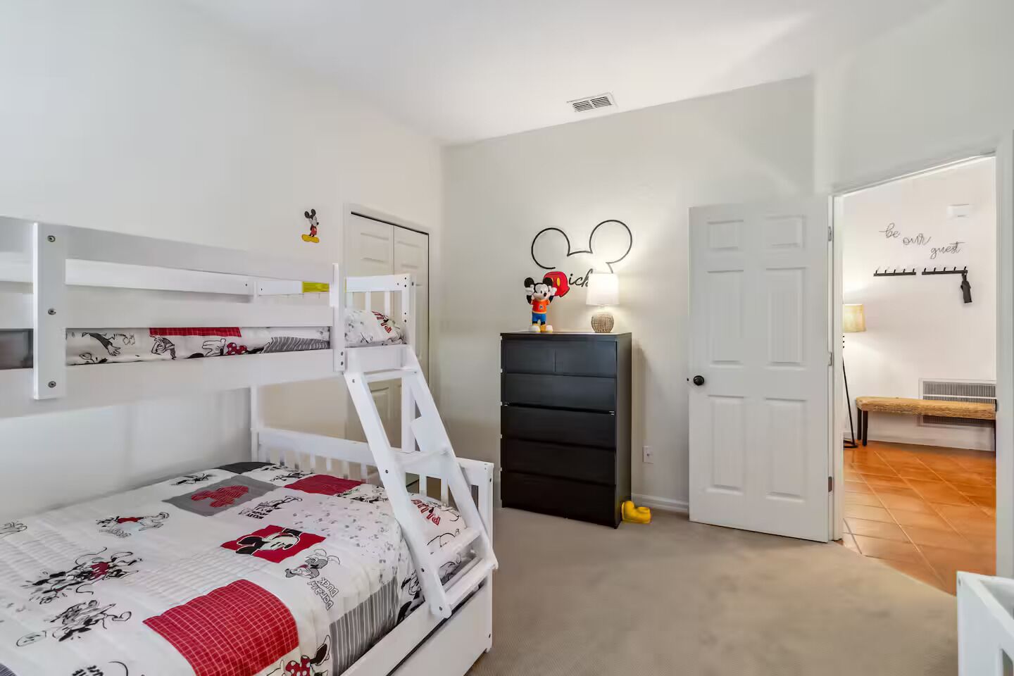 Quarto com cama de solteiro e beliche, cômoda, decoração de parede com tema do Mickey Mouse e porta com uma placa.
