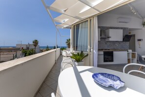 A balcony with a view of the sea, a dining area with a white table and chairs, and a kitchen with white cabinets and a tiled backsplash. - Apartment 'Attico' with Sea View, Wi-Fi and Air Conditioning (Donnalucata)