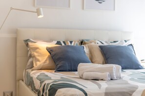 Una cama bien hecha con una colcha estampada, una pila de toallas blancas y una lámpara de noche.