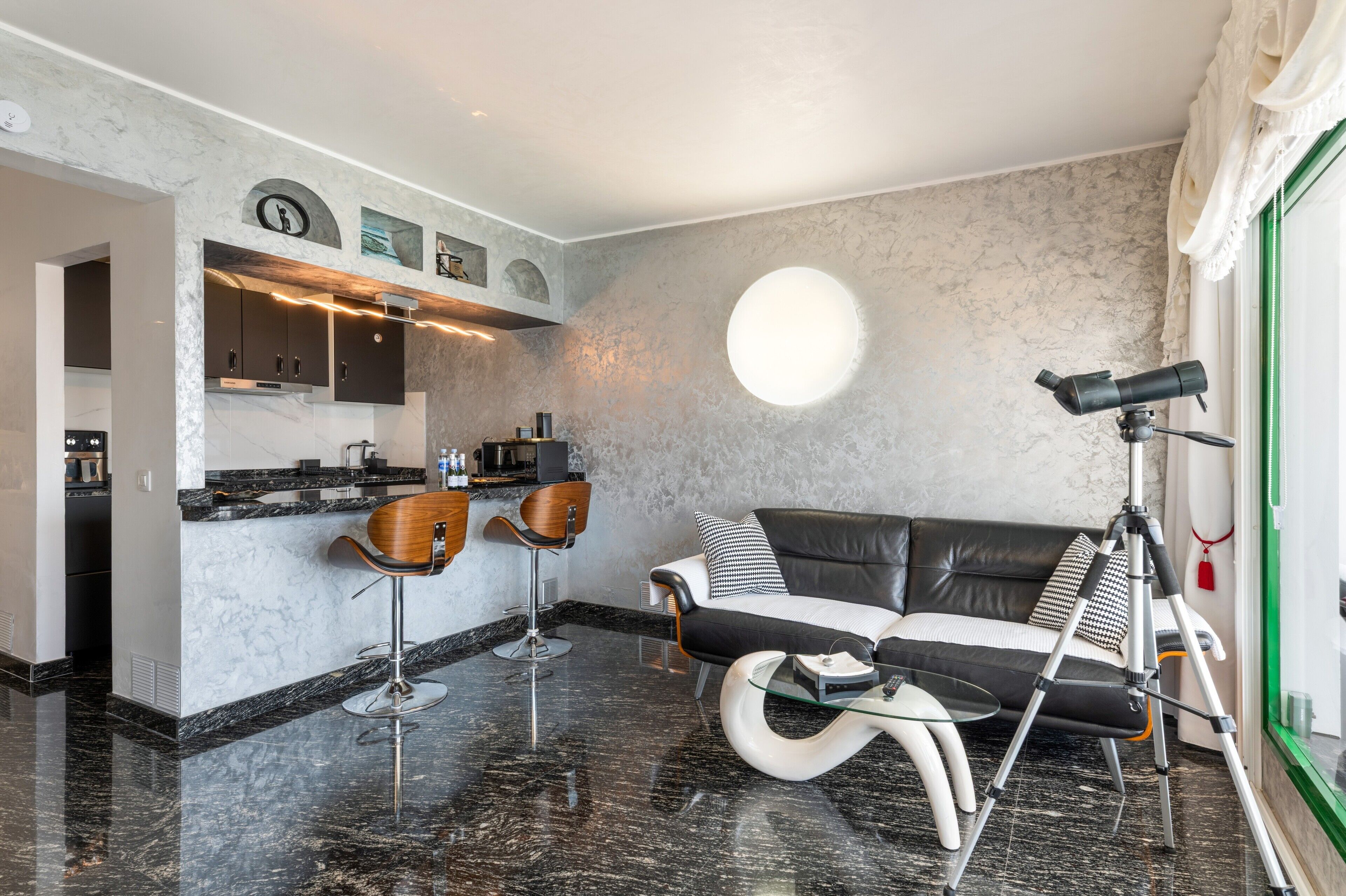 A modern living room with a black leather sofa, a glass coffee table, and a bar area with stools.