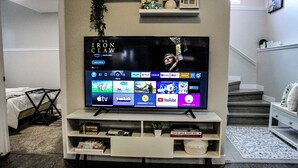 A living room with a flat-screen TV displaying a movie, a bookshelf with books and decorative items, a coffee table with a plant, and a staircase in the background. - Home away from home (Grande Prairie)