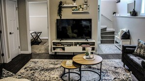 A living room with a TV, a coffee table, and a sofa. There is a staircase leading to another level. - Home away from home (Grande Prairie)
