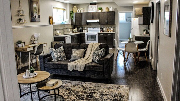 A compact living space with a dark-colored sofa, a small round table, and a kitchen area with dark wooden cabinets and a white stove. - Home away from home (Grande Prairie)