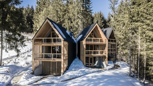 Twee houten hutten met grote ramen, omgeven door sneeuw en bomen.