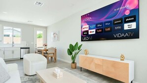 A modern living room with a flat-screen TV displaying various streaming apps, a white sofa, a wooden sideboard, and a dining area in the background. - 3BD / Free Parking / 75" Smart TV / 2BR / 2Bath (Glendale)