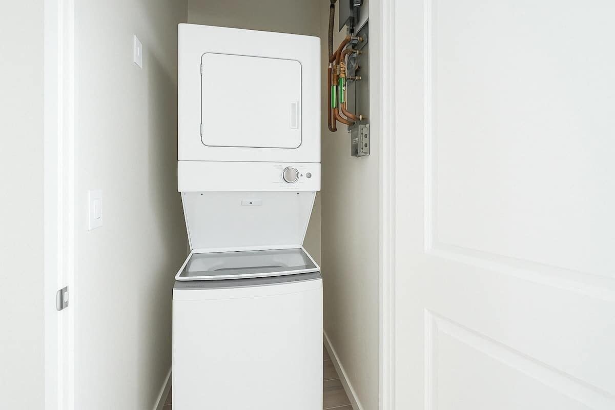 Een kleine, witte wasruimte met een ingebouwde wasmachine en droger, een bedieningspaneel en zichtbare leidingen.