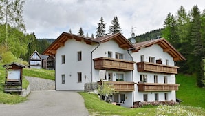 A two-story chalet with a balcony, surrounded by a lush green landscape and a clear sky.
