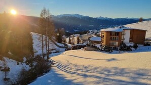 Schneebedeckte Landschaft mit einem Gebäude, das eine Holzfassade und ein geneigtes Dach hat, umgeben von Bergen und Bäumen.