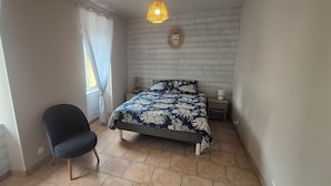 A bedroom with a bed, a chair, a bedside table, a lamp, and a window with curtains.