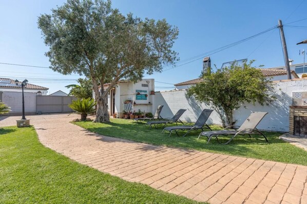 A paved pathway leading to a house with a white exterior, a tree, and two lounge chairs.