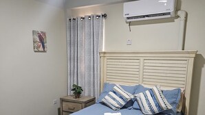 Ein ordentlich bezogenes Bett mit blauer Bettdecke und Kissen, ein kleiner Nachttisch mit Pflanze und eine Klimaanlage über dem Bett.
