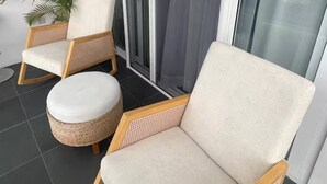 Ein Balkon mit zwei Korbstühlen, einem runden Hocker, einem Blumentopf und einer Glastür, die auf eine Terrasse führt.